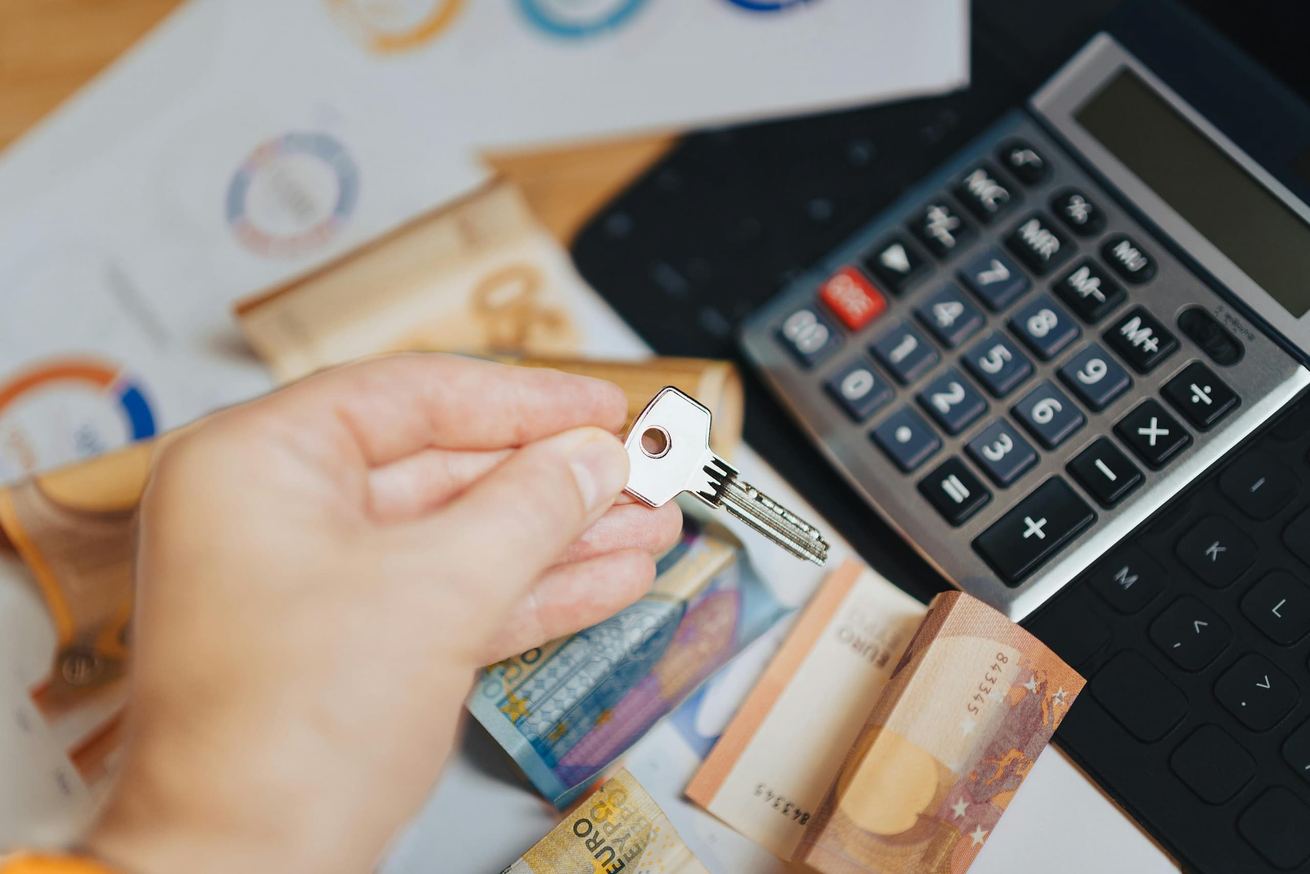 A close-up of a hand holding a key above a table with Euro banknotes, a calculator, and documents, symbolizing financial planning for down payment assistance programs for first-time homebuyers.