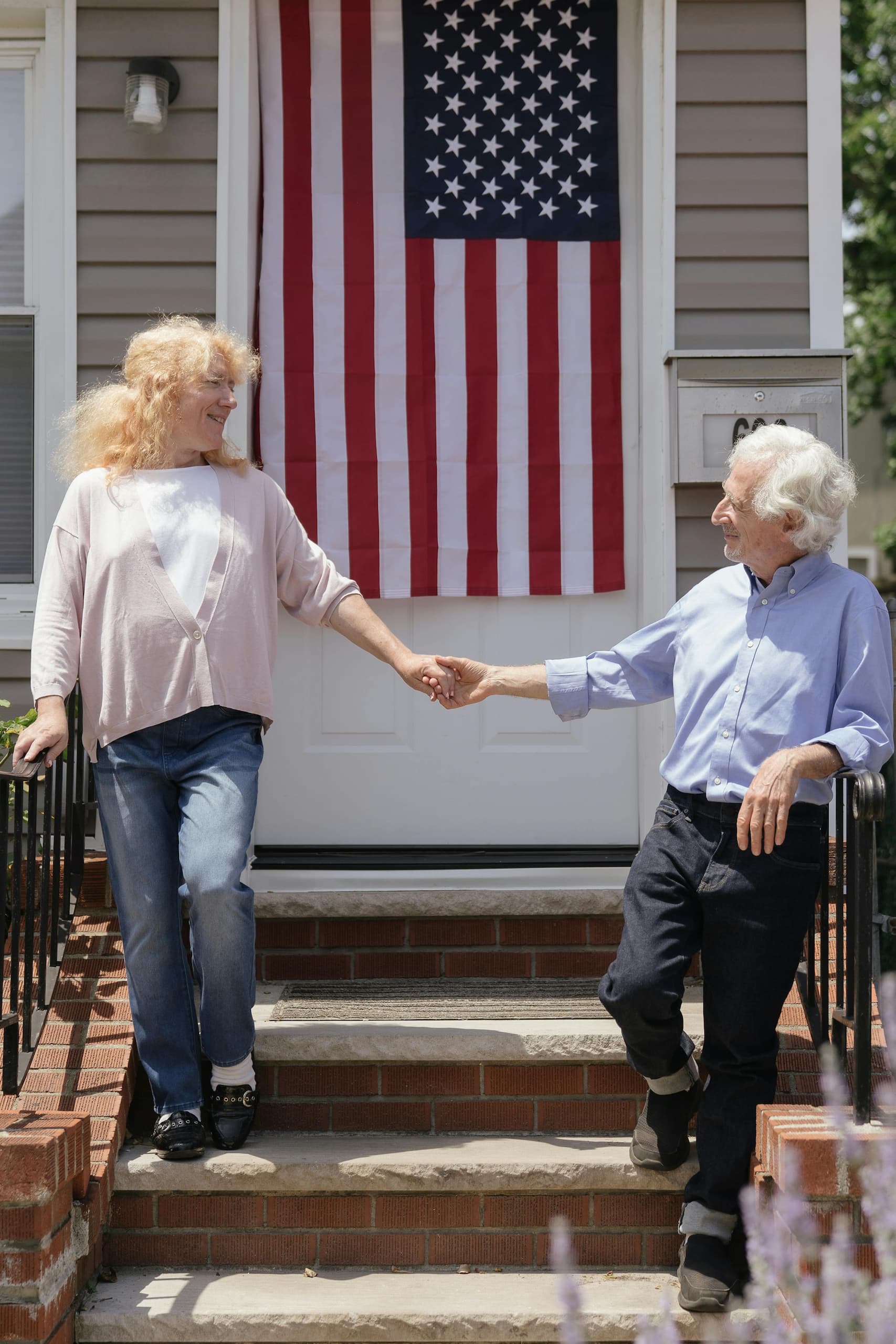 Elderly couple holding hands on the steps of their home, with an American flag in the background, symbolizing homeownership opportunities for veterans through VA Home Loans